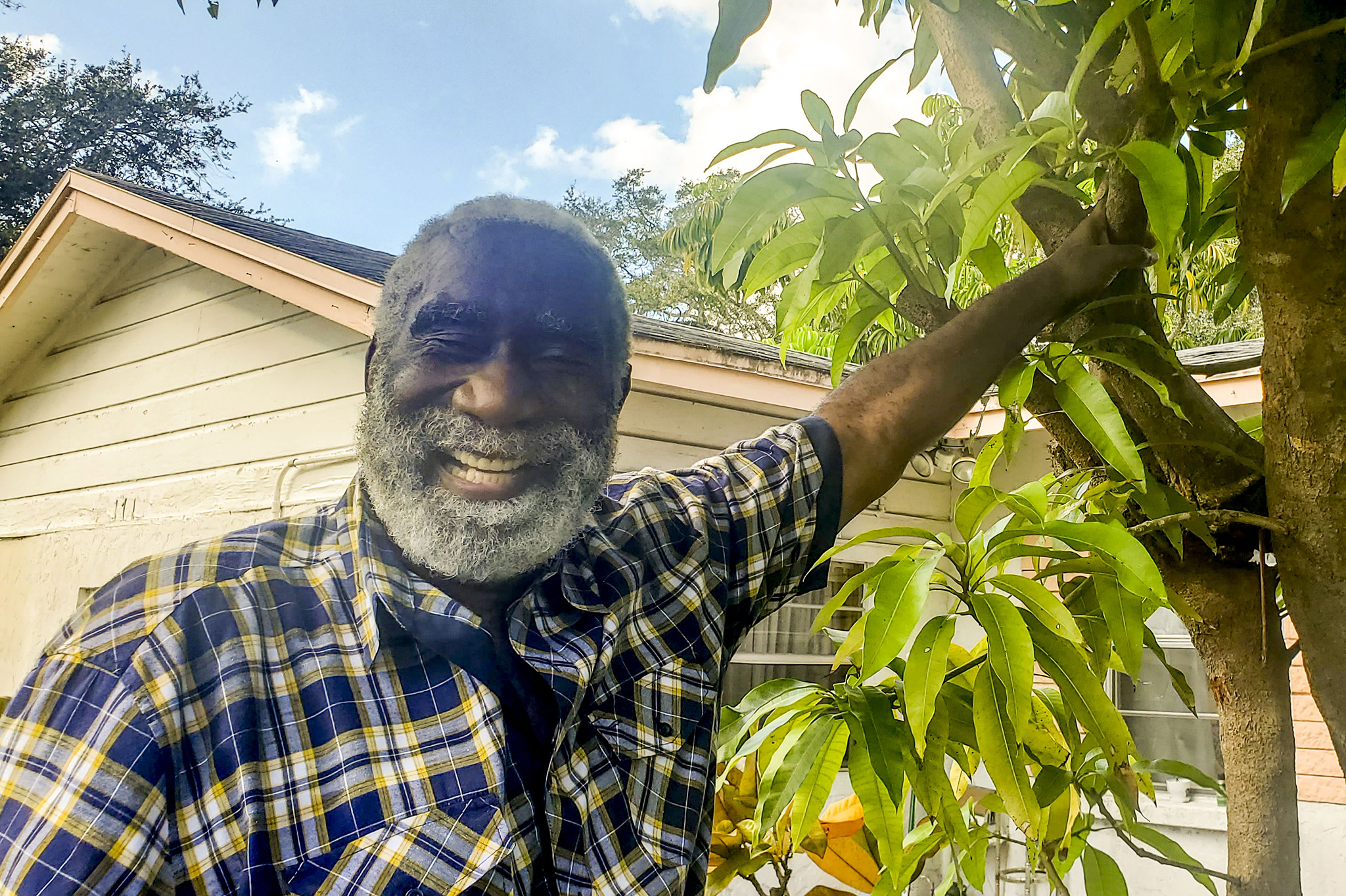 Fredd Atkins, an older African-American man, stands smiling with his hand resting on a mango tree in front of a house. It is a sunny day and blue skies can be seen in the background.