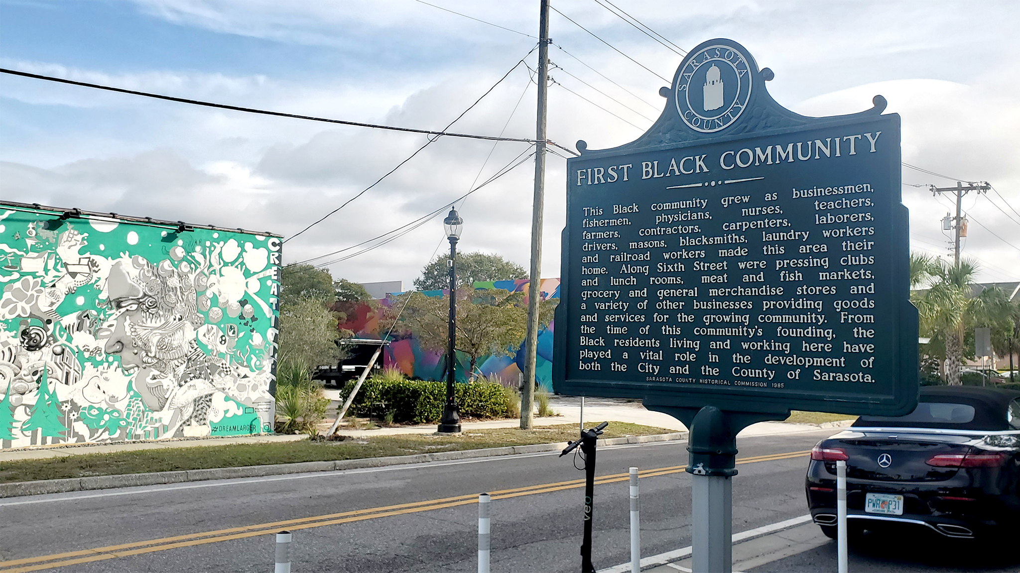 A black plaque with gold lettering titled “First Black Community” that reads “This Black community grew as businessmen, fishermen, physicians, nurses, teachers, farmers, contractors carpenters, laborers, drivers, masons, blacksmiths, laundry workers and railroad workers made this area their home. Along Sixth Street were pressing clubs and lunch rooms, meat and fish markets, grocery and general merchandise stores and a variety of other businesses providing goods and services for the growing community. From the time of this community’s founding, the Black residents living and working here played a vital role in the development of both the City and the County of Sarasota.”